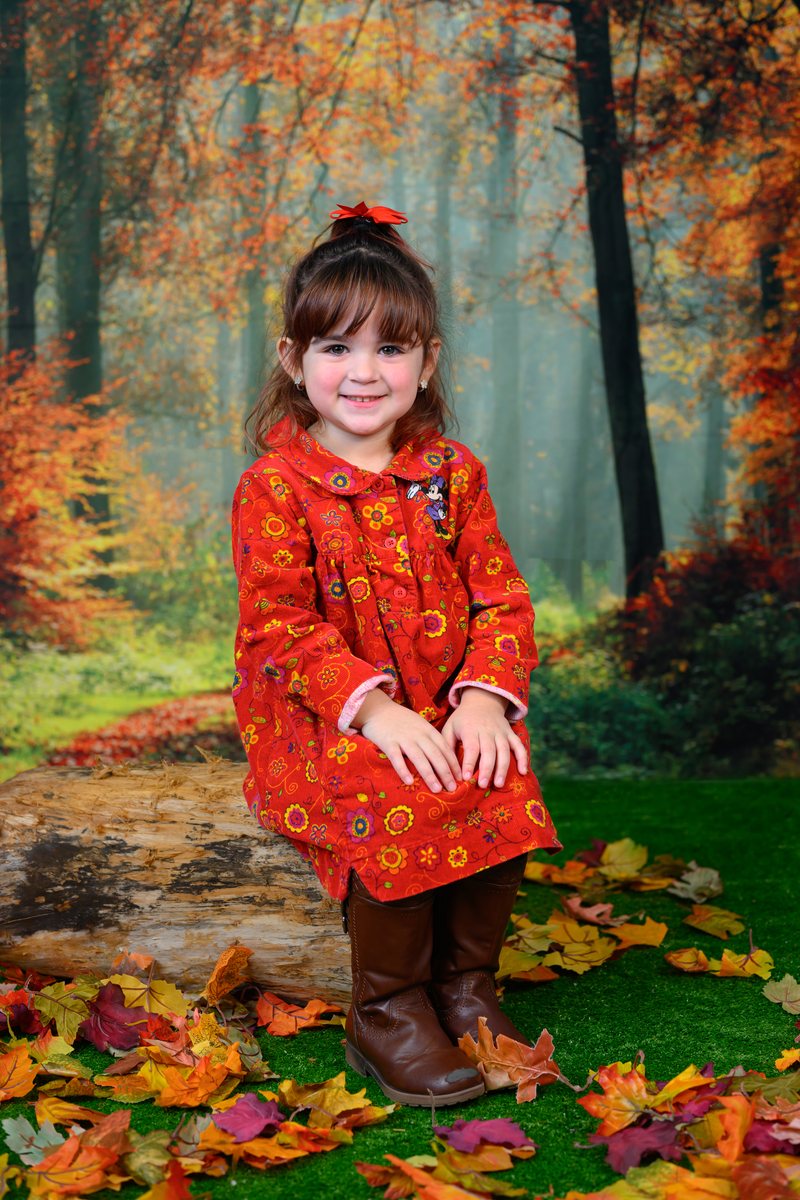 Girl in a red flower dress smiling, seated on a log in an autumn scene